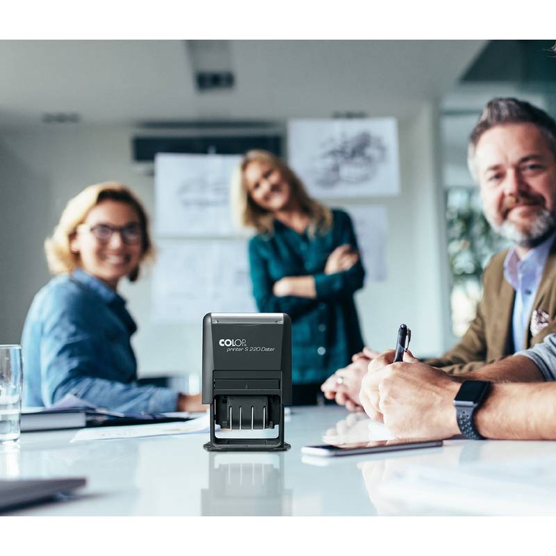 Bürobesprechung mit vier lächelnden Personen an einem Tisch mit Dokumenten; ein schwarzer Datumsstempel ist auf der Tischoberfläche im Fokus.