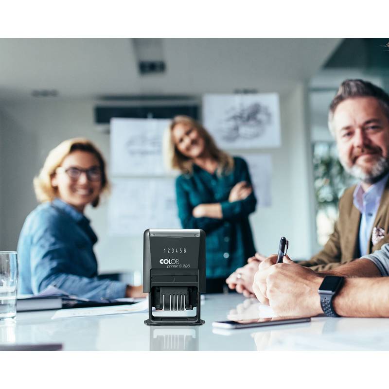 Eine Gruppe von Menschen in einem Besprechungsraum, die lächelnd in die Kamera blicken, mit Fokus auf einem Datumsstempel in der Mitte, was eine kollaborative Atmosphäre andeutet.