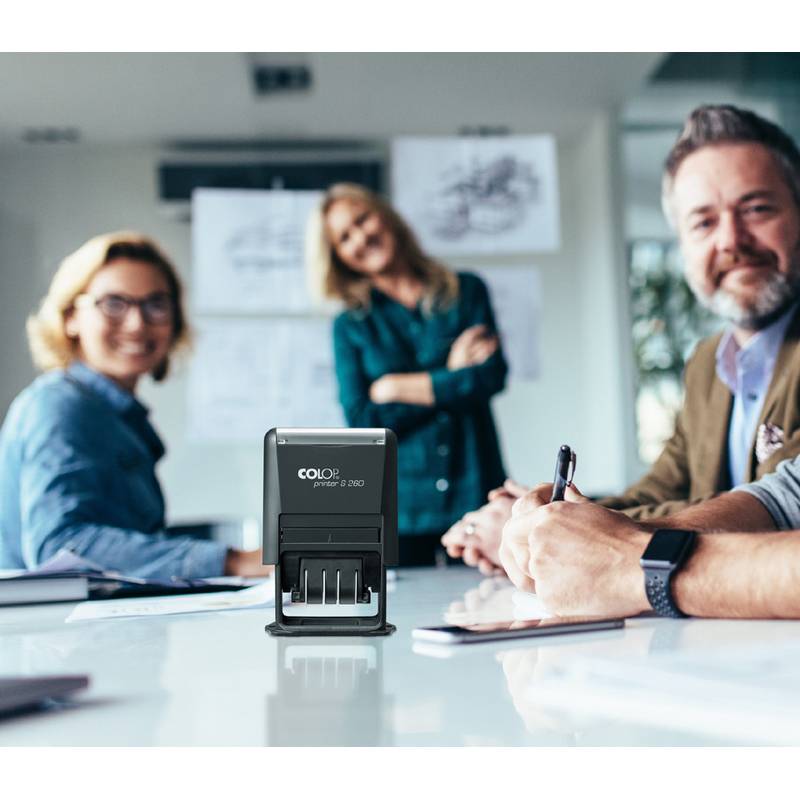 Eine Gruppe von Menschen, die um einen Konferenztisch sitzen, lächeln und Papiere sowie einen COLOP-Stempel auf dem Tisch haben, was eine Besprechungssituation andeutet.