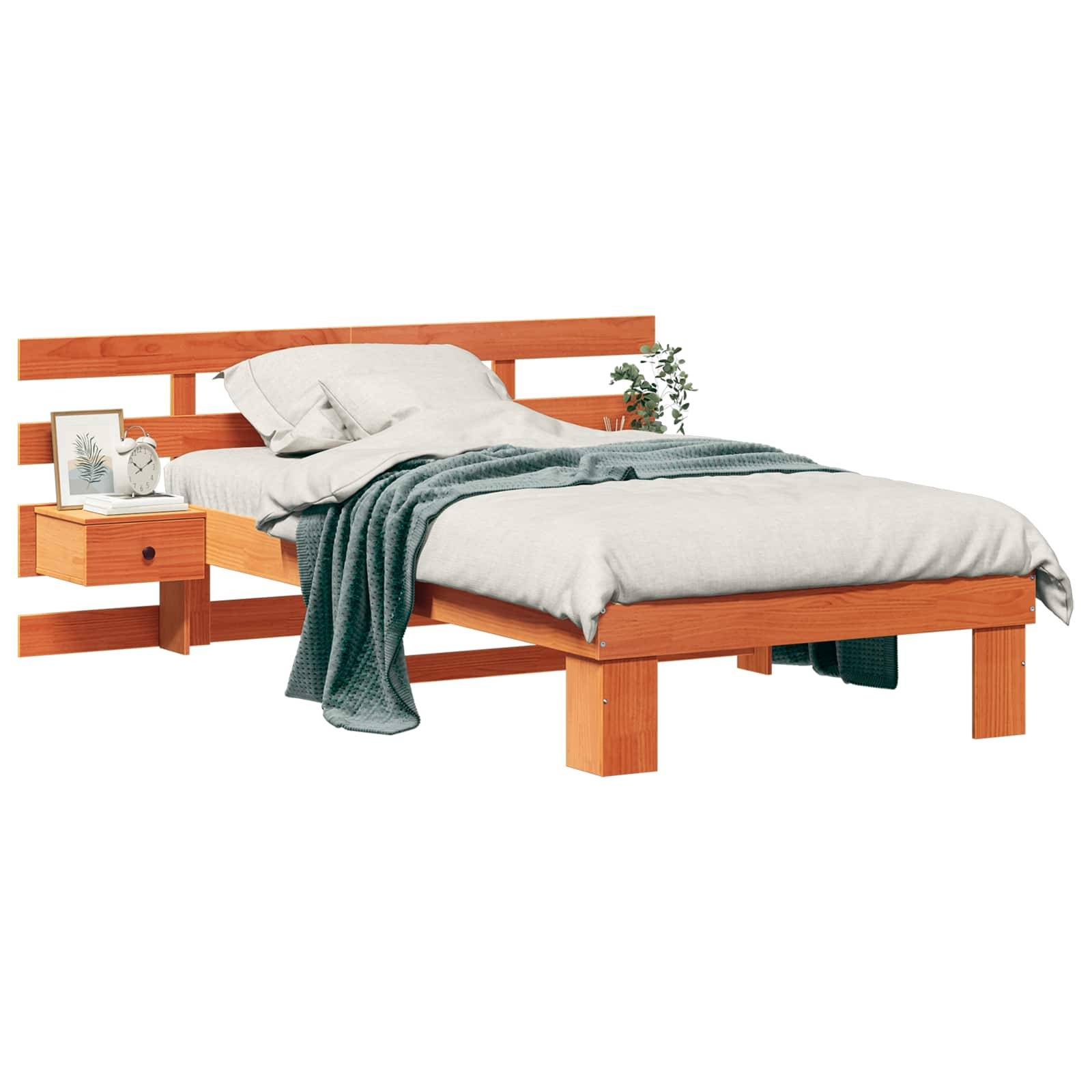 Ein Holzbett mit einer hellbeigen Bettdecke und Kissen, eine grüne Decke, die seitlich darüber drapiert ist. Ein Nachttisch trägt einen Bilderrahmen und eine kleine Pflanze.