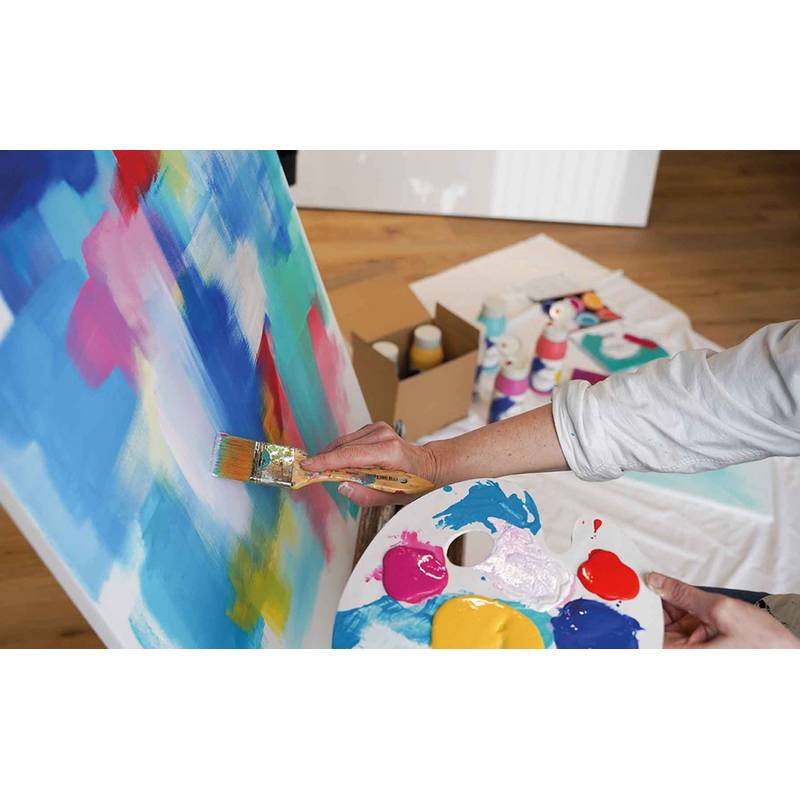 Eine Person malt eine farbenprächtige abstrakte Leinwand mit einem Pinsel und einer Palette mit verschiedenen Farben. Malutensilien sind auf dem Boden verstreut.