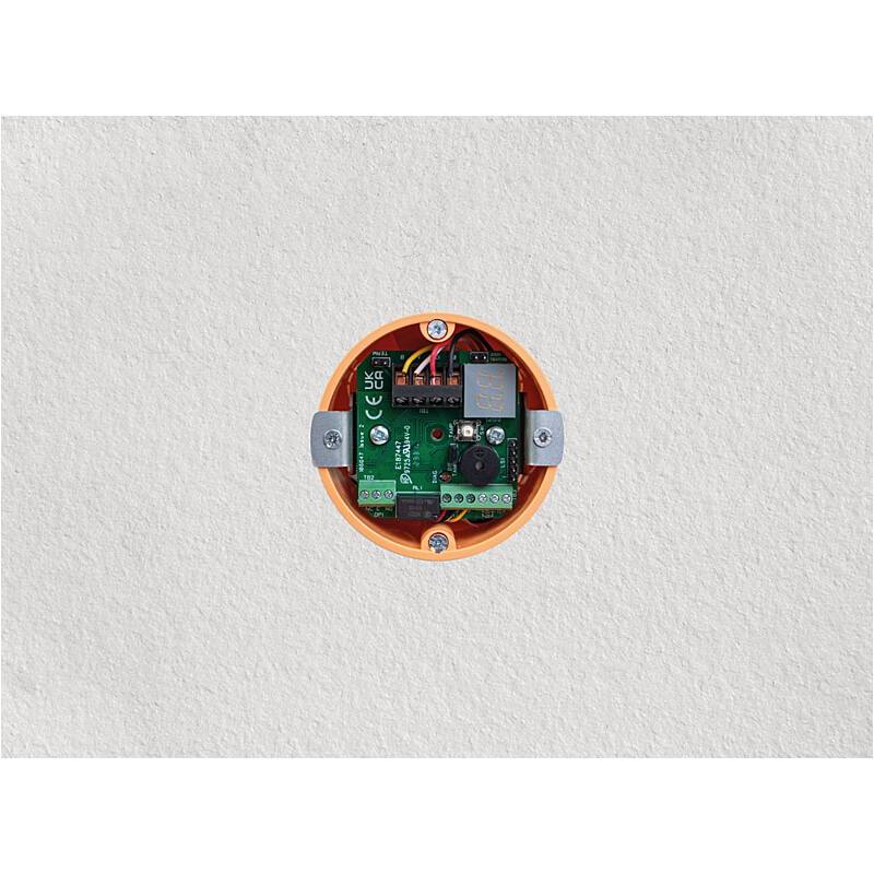 Eine kleine elektrische Schalttafel, die in einer runden orangefarbenen Wanddose an einer einfachen weißen Wand angebracht ist.