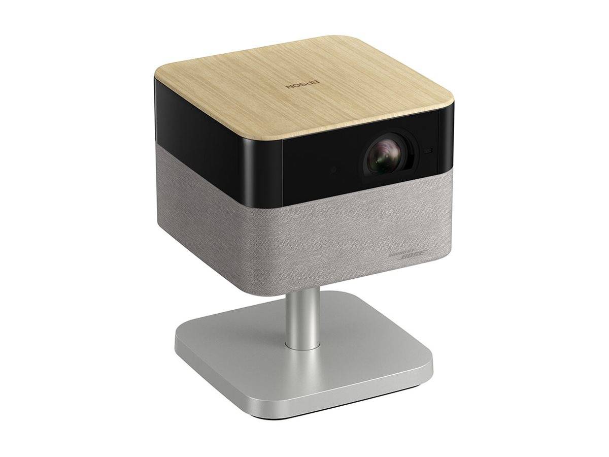 Ein kompakter, moderner Projektor mit einer Holzoberfläche auf der Oberseite und stoffbezogenen Seitenflächen, montiert auf einem schlanken Metallständer.