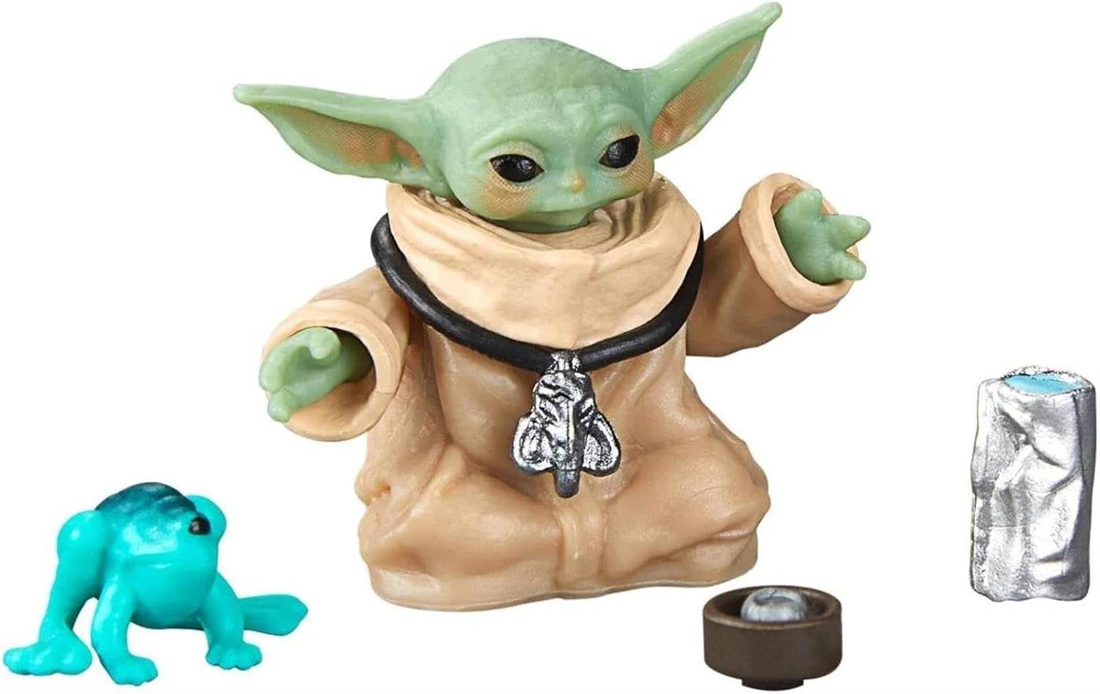 Eine Baby-Yoda-Figur mit einer Halskette, gezeigt mit einem kleinen blauen Frosch, einer Tasse und einem silbernen Behälter auf weißem Hintergrund.