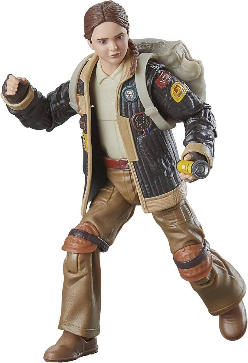 Actionfigur einer jungen Frau in einer Jacke und beigefarbenen Hosen, in einer dynamischen Pose mit einer erhobenen Faust und einem Gerät in der anderen Hand.