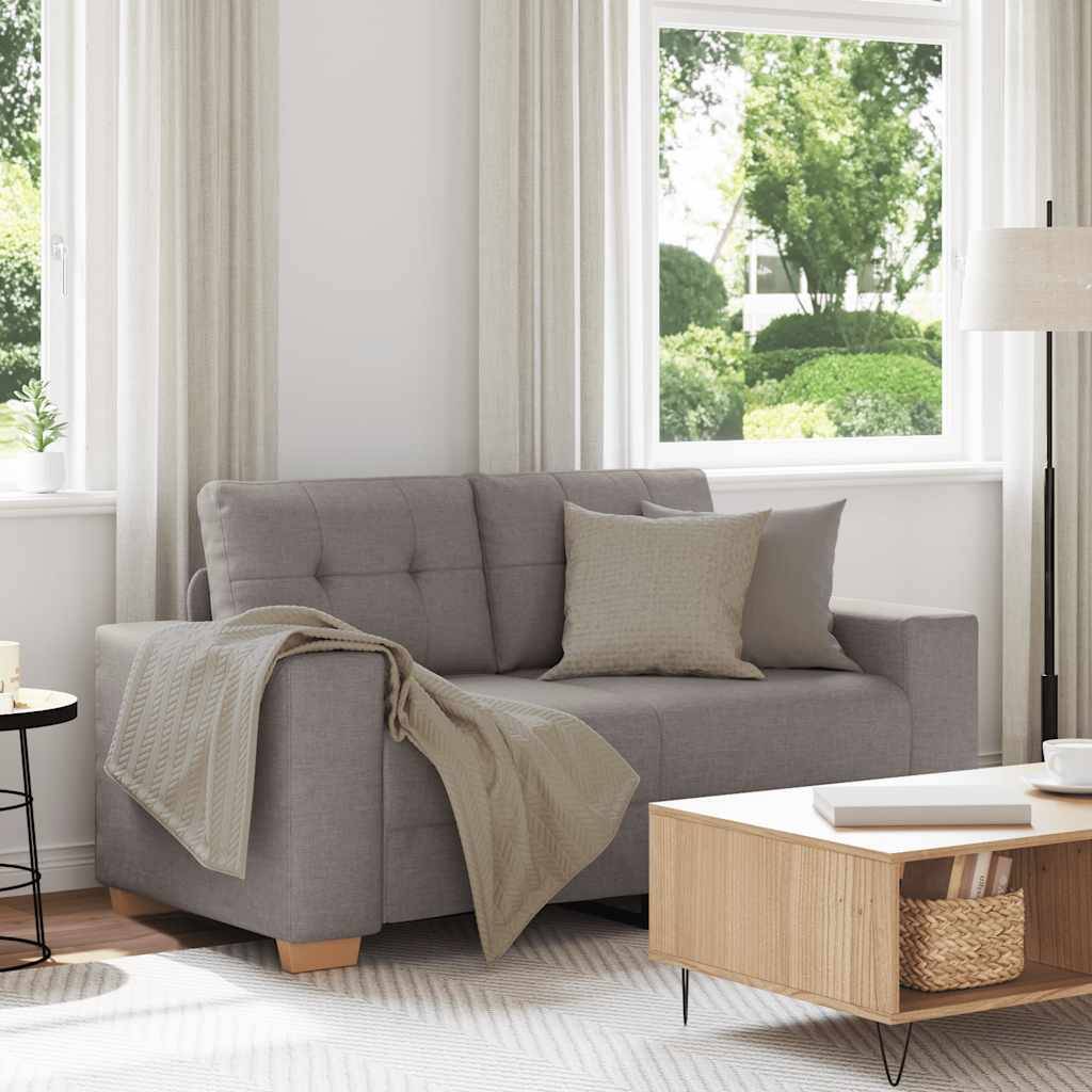 Ein modernes Wohnzimmer mit einem grauen Sofa, beigen Kissen und einer Decke. Ein hölzerner Couchtisch trägt ein Buch. Große Fenster zeigen grüne Bäume draußen.