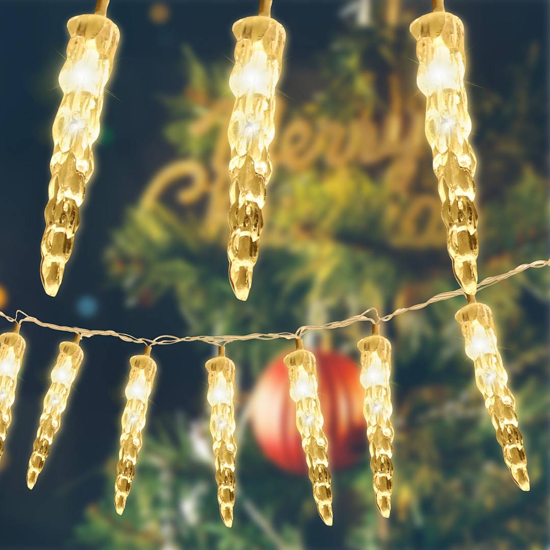 Warmweiße Eiszapfen-förmige Lichterketten hängen vor einem unscharfen Weihnachtsbaum mit Christbaumkugeln, was auf weihnachtliche Dekoration hindeutet.