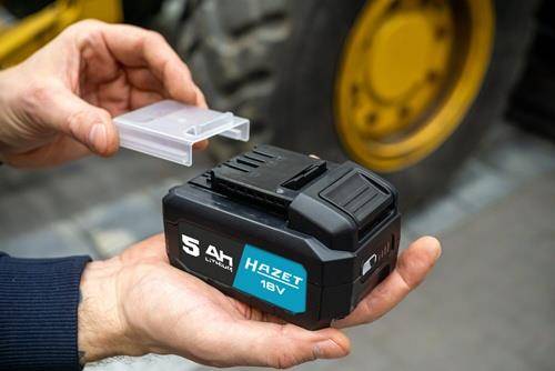 Eine Person hält ein aufladbares 'Hazet 18V 5Ah Li-Ion' Akkupack, während sie dessen Plastikabdeckung entfernt, mit verschwommener Maschinerie im Hintergrund.