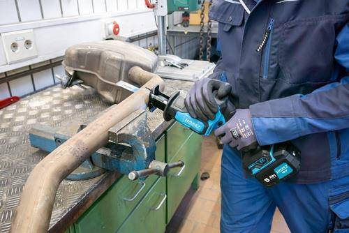 Eine Person mit Schutzhandschuhen verwendet eine tragbare Kreissäge, um ein Metallrohr zu durchtrennen, das auf einer Werkbank in einer Werkstattumgebung eingespannt ist.