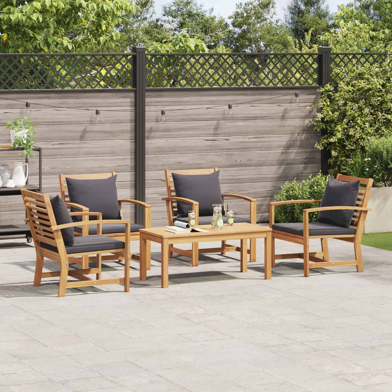 Vier hölzerne Gartenstühle mit dunklen Polstern sind um einen passenden Holztisch auf einer Terrasse angeordnet, mit Pflanzen und einem Holzzaun im Hintergrund.