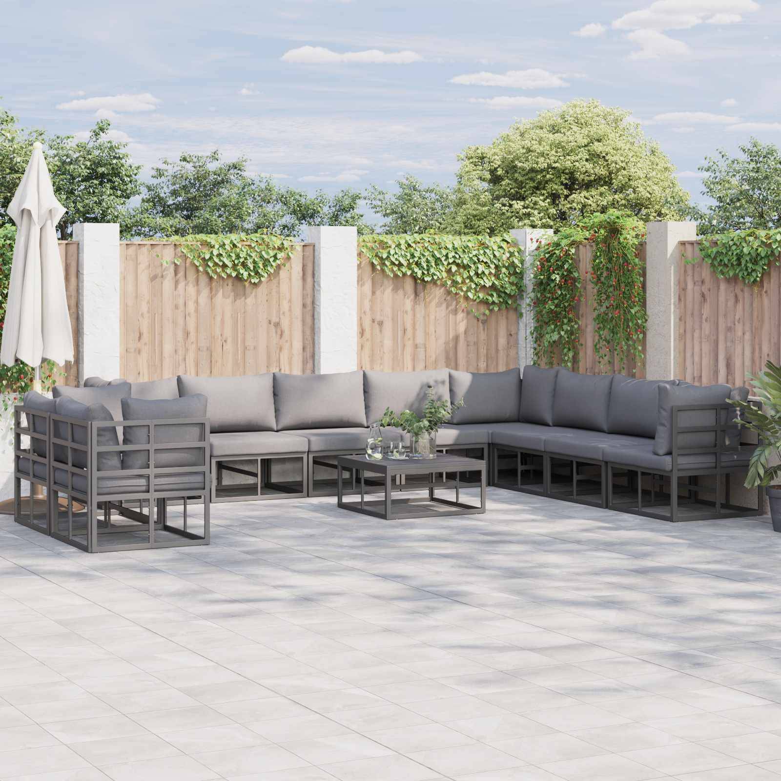 Terrasse mit grauen Ecksofa, einem Couchtisch und umgebendem Holzzaun vor einem Hintergrund von Grünflächen und blauem Himmel.
