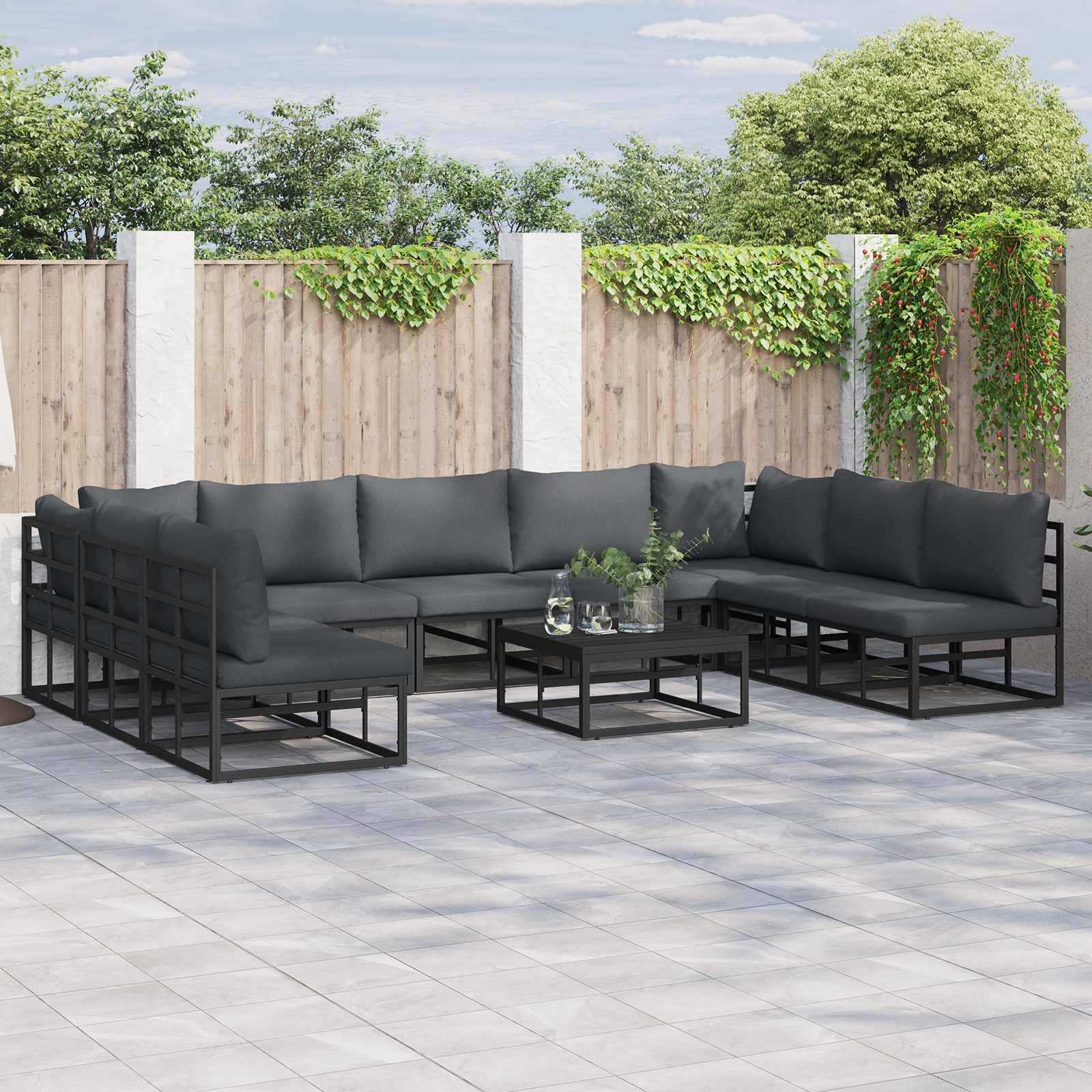 Überdachte Terrasse mit einem dunkelgrauen Ecksofa-Set auf Bodenfliesen, umgeben von einem Holzzaun mit Grünpflanzen und klarem Himmel.