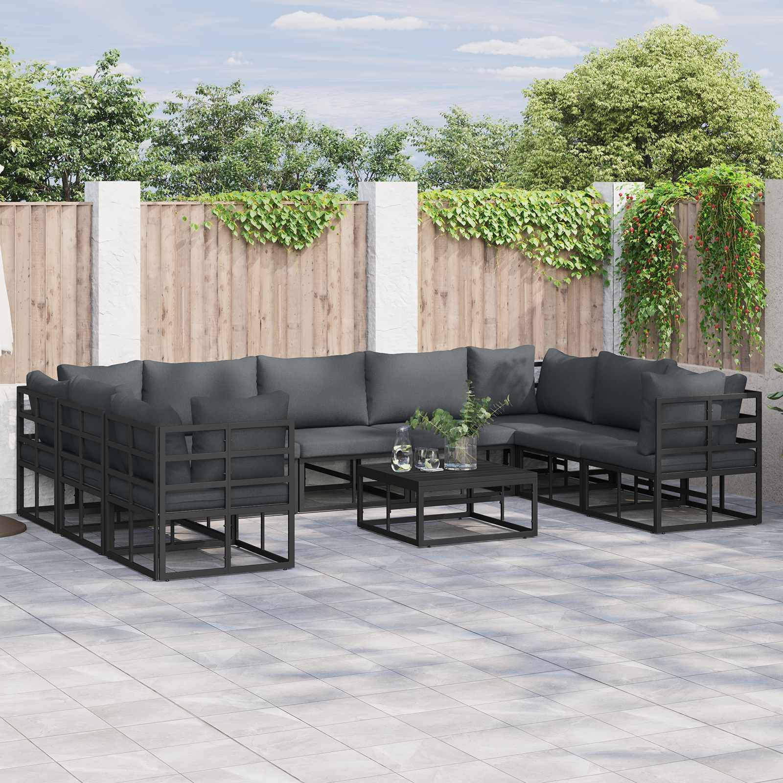 Outdoor-Terrasse mit grauem Ecksofa und Tisch auf gefliesten Boden, umgeben von einem Holzzaun, über den Grünpflanzen klettern.