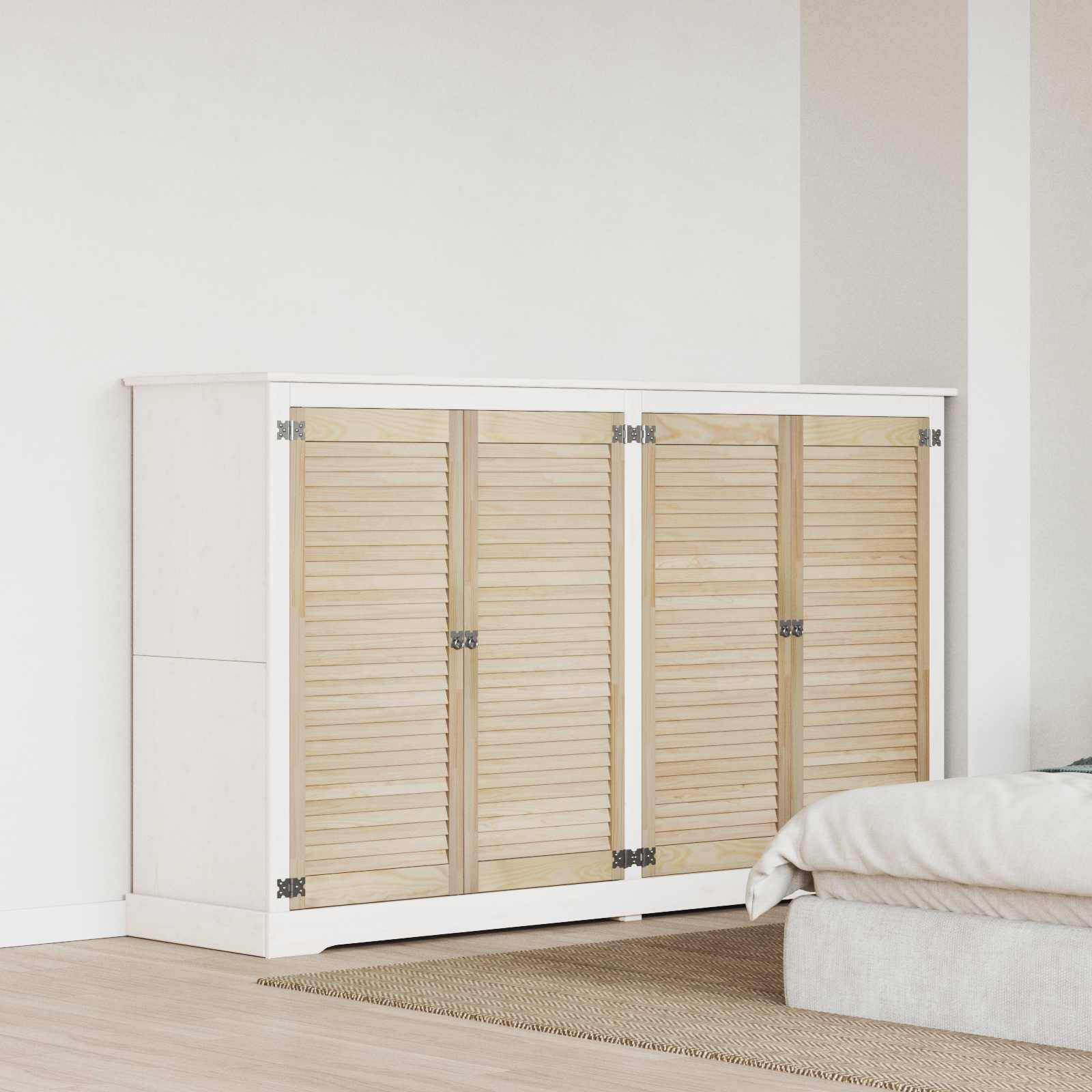 Ein Holzschrank mit vier Lamellentüren steht an einer weißen Wand auf einem beigen Teppich, neben einem Stapel hellgefärbter Kissen.