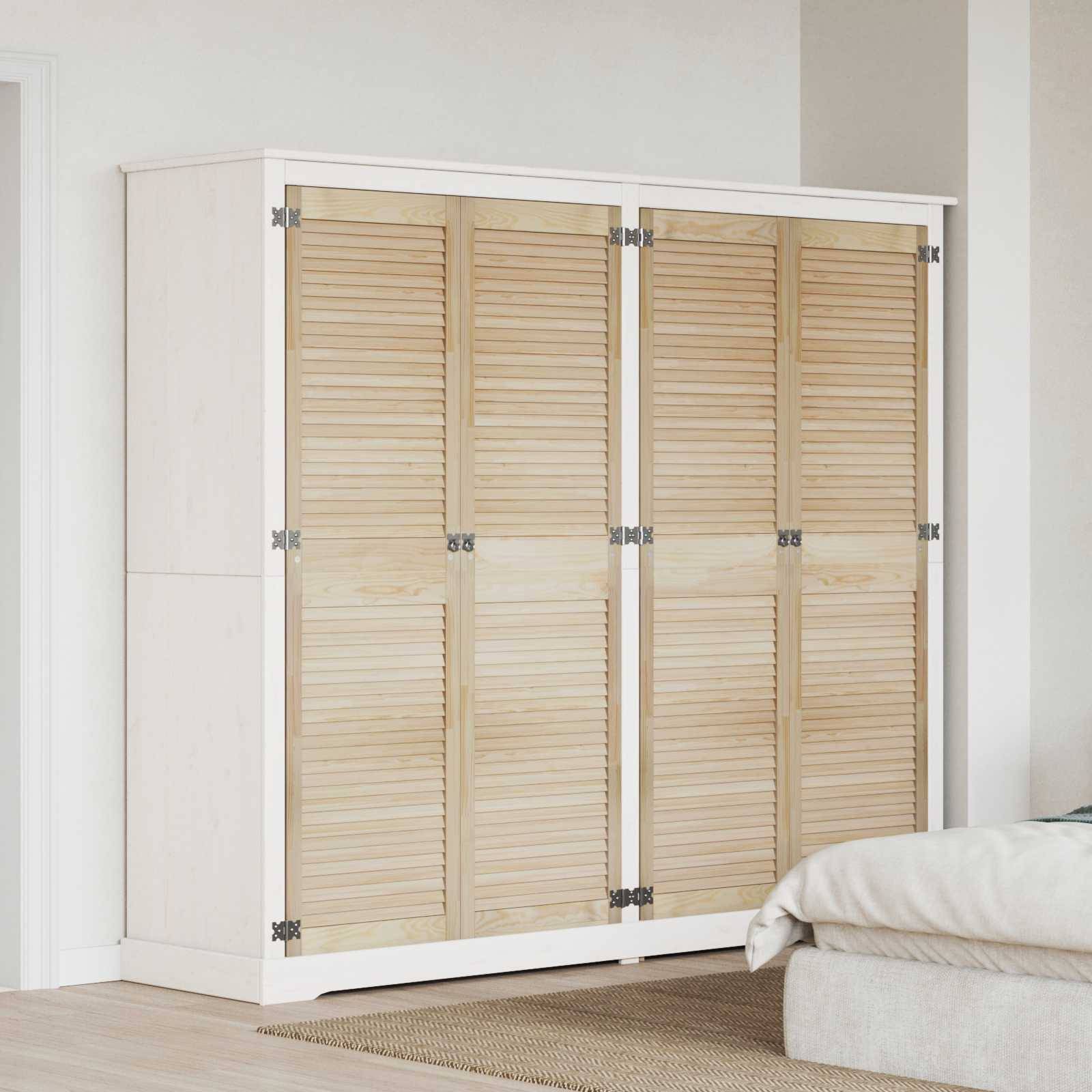Ein weißer Kleiderschrank mit Lamellentüren aus Holz steht neben einer schlichten Wand. Ein beiges Bett, das rechts teilweise zu sehen ist, deutet auf eine Schlafzimmerumgebung hin.