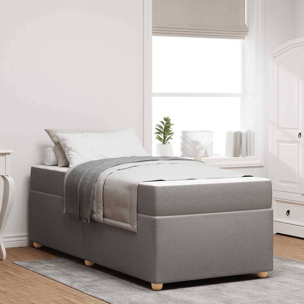 Ein ordentlich gemachtes grau gepolstertes Bett mit Kissen und einer weiß-grauen Steppdecke steht in einem hellen Raum mit großen Fenstern und Büchern auf einem Regal.