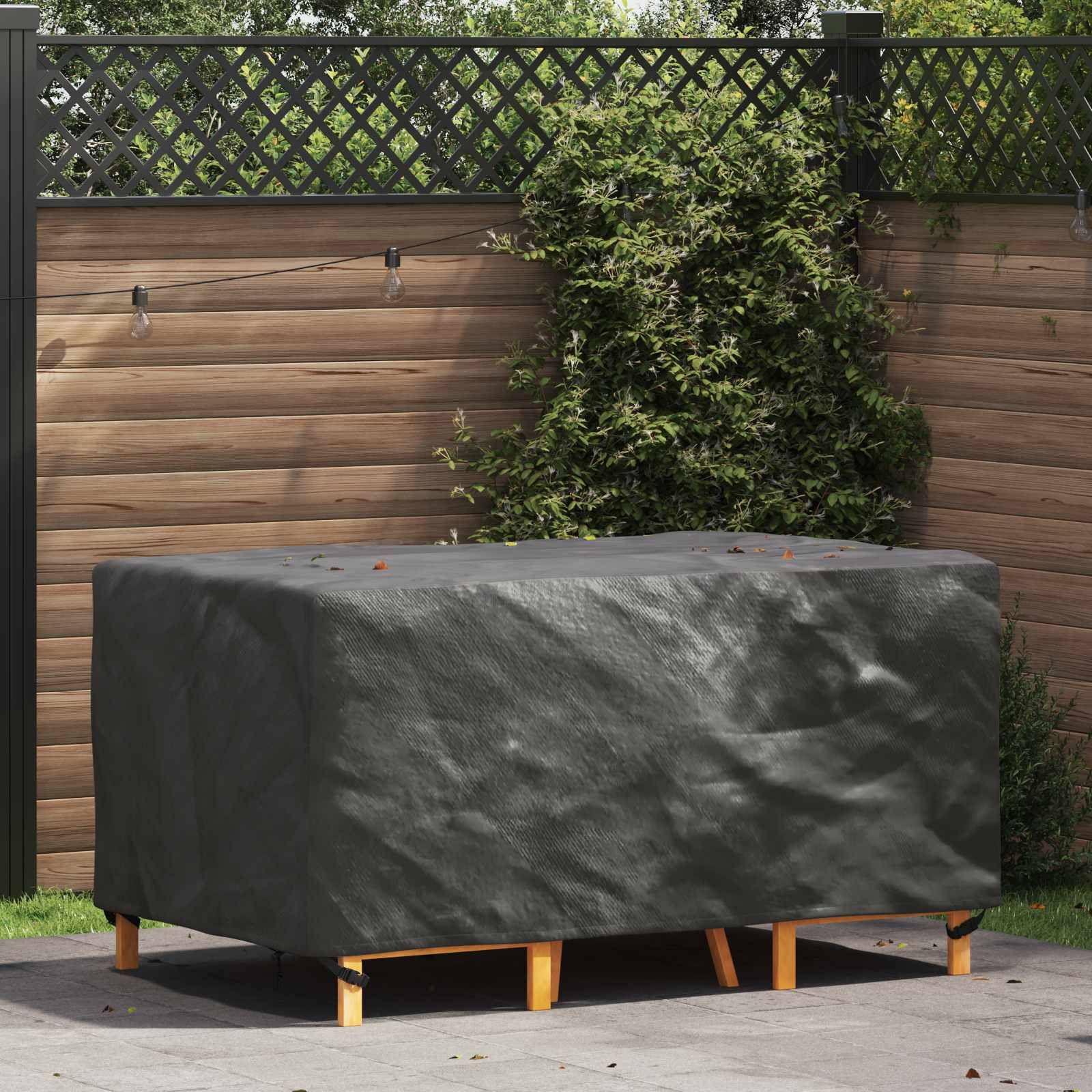 Gartentisch mit einer grauen Schutzhülle in einer Gartenumgebung, umgeben von Grünpflanzen und einem Gitterzaun mit Lichterketten.