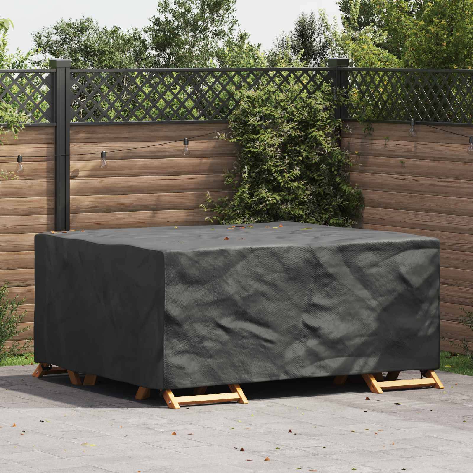 Ein rechteckiger Outdoor-Tisch, der mit einer schwarzen Schutzabdeckung versehen ist, steht auf einer Terrasse, umgeben von einem Holzzaun und üppigem grünem Blattwerk.