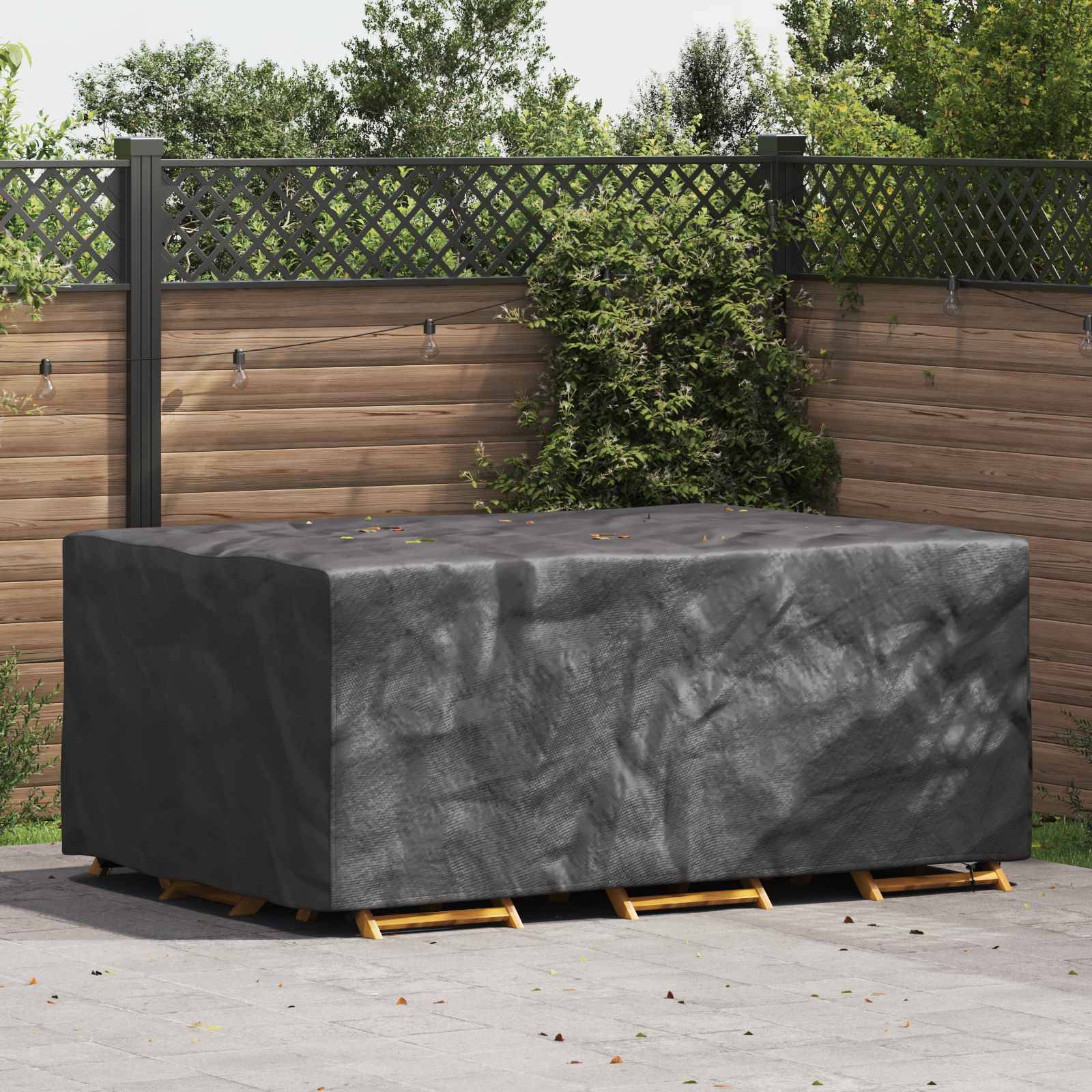 Ein großer rechteckiger Outdoor-Tisch, der mit einer dunkelgrauen Schutzabdeckung versehen ist, auf einer Terrasse mit Holzzaun und grünem Blattwerk im Hintergrund.