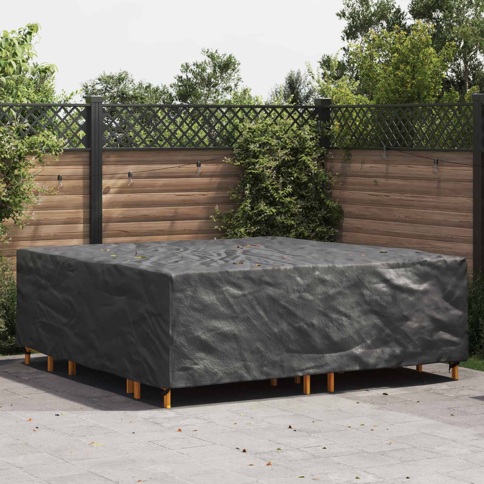 Ein großer Outdoor-Esstisch ist mit einer dunklen Schutzplane bedeckt, aufgestellt auf einer gepflasterten Fläche, umgeben von einem Holzzaun und Grünbewuchs.