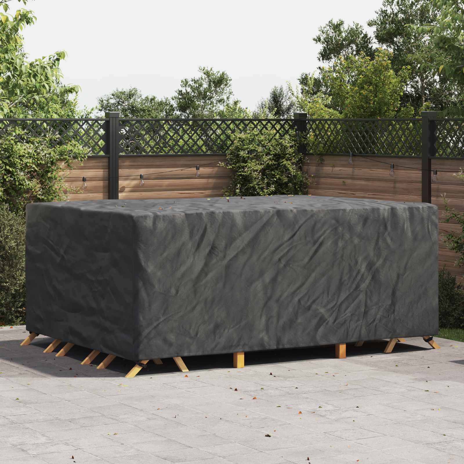 Ein großer Outdoor-Tisch ist mit einem dunklen, schützenden Stoff bedeckt. Er steht auf einer Terrasse, umgeben von Holzzaun und grünem Blattwerk.