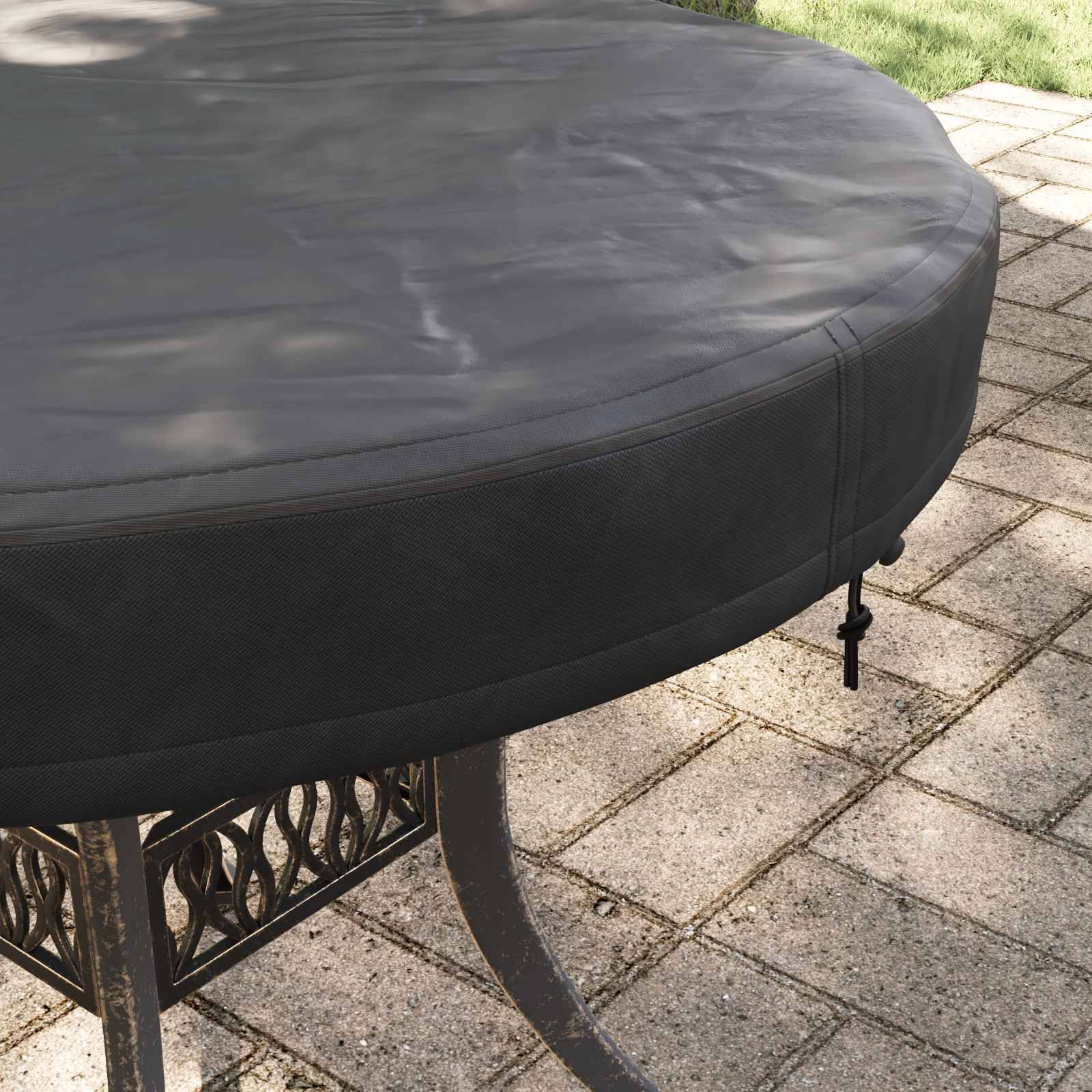 Ein runder Terrassentisch, der mit einer passgenau anliegenden, grauen Schutzhülle bedeckt ist, steht auf einer mit Natursteinen gepflasterten Terrasse, umgeben von grünem Rasen.