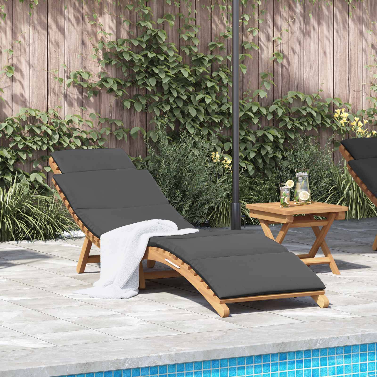 Zwei Holzliegen mit dunklen Kissen stehen neben einem Pool. Auf einem kleinen Tisch stehen ein Krug und Becher. Im Hintergrund befindet sich ein Zaun mit Blattwerk.
