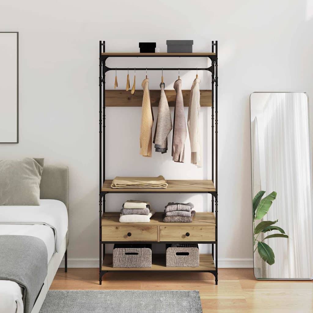 Ein Holzschrank mit hängenden Kleidungsstücken und Regalen, auf denen gefaltete Kleidung und Aufbewahrungsboxen liegen. Er steht neben einem Bett und einem hohen Spiegel.
