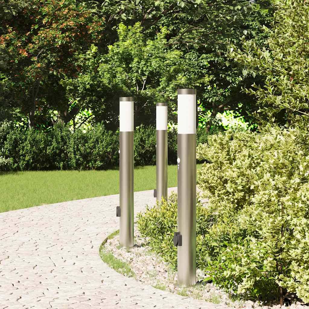Ein Gartenweg ist gesäumt von modernen, hohen, silbernen Laternen, die von üppigem Grün umgeben sind. Zurückspulen