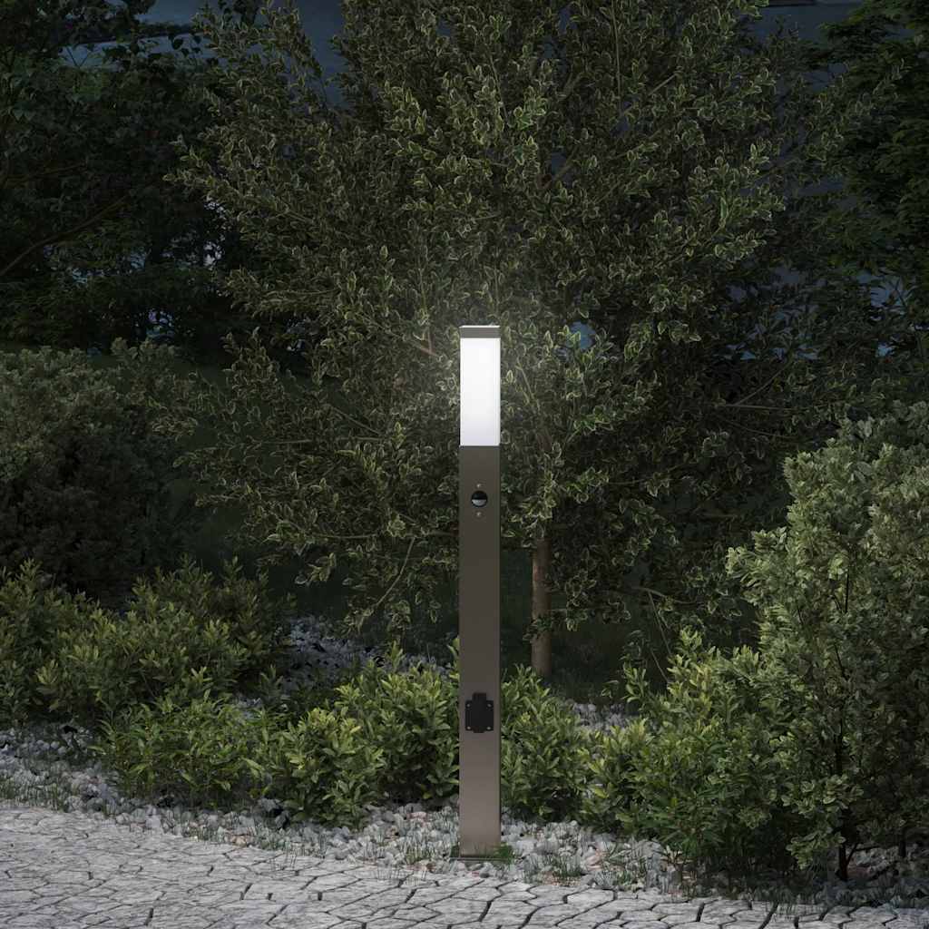 Eine moderne Gartenwegleuchte steht zwischen dichtem Grün in der Abenddämmerung und beleuchtet einen Kopfsteinpflasterweg sowie die umgebenden Sträucher.