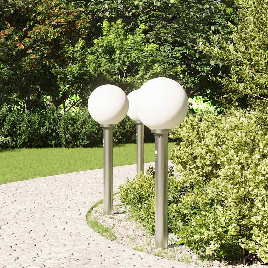 Ein geschwungener Steinweg in einem Garten mit Gras und Büschen, auf dem drei weiße kugelförmige Lampen auf Metallständern stehen, umgeben von Grünpflanzen.