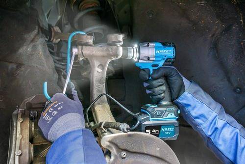 Eine Person verwendet einen elektrischen Schlagschrauber, um eine Schraube an einer Fahrzeugaufhängung zu befestigen. Blaue und schwarze Handschuhe und Werkzeuge sind sichtbar.