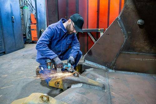 Ein Arbeiter in blauen Overalls und einer Kappe benutzt einen Winkelschleifer an einer Metallkonstruktion und erzeugt dabei Funken in einer industriellen Umgebung.