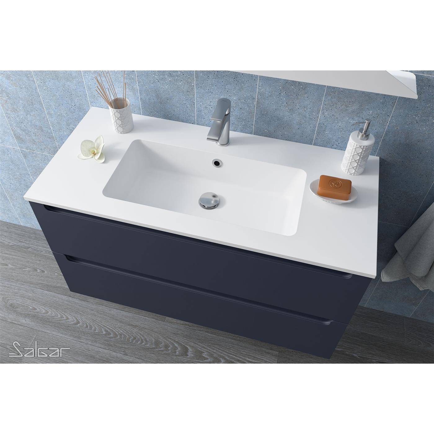 Modernes Badezimmerwaschtisch mit weißer Arbeitsplatte und Waschbecken, Chromarmatur, dunklem Unterschrank und kleinen Accessoires vor blauer Fliesenwand.