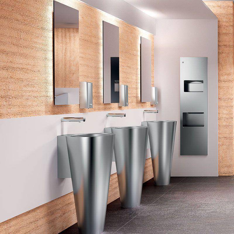 Modernes öffentliches Badezimmer mit drei Edelstahl-Säulenbecken, verspiegelten Stationen und einem wandmontierten Händetrockner oder Spender auf der rechten Seite.
