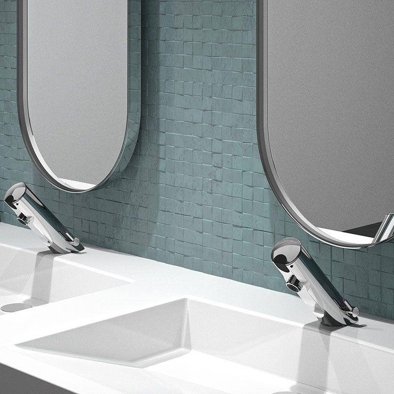 Modernes Badezimmer mit zwei verchromten automatischen Wasserhähnen über einem weißen Doppelwaschbecken, umrahmt von ovalen Spiegeln und einer blaugrün gefliesten Wand.