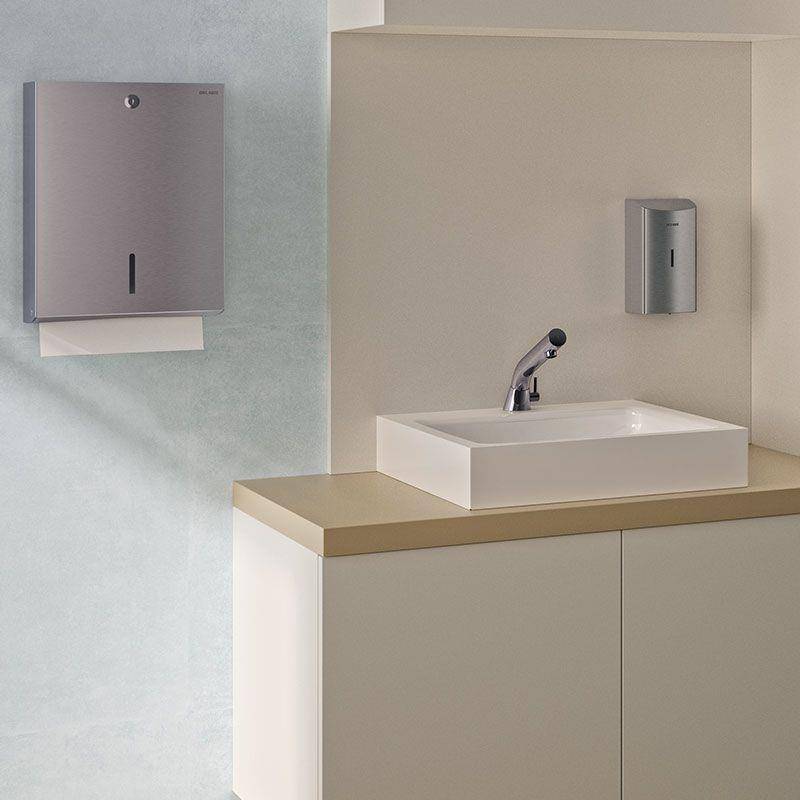 Minimalistisches Badezimmerbecken mit wandmontiertem Waschbecken, Armatur und zwei Edelstahl-Spendern an einer beige und blauen Wand.