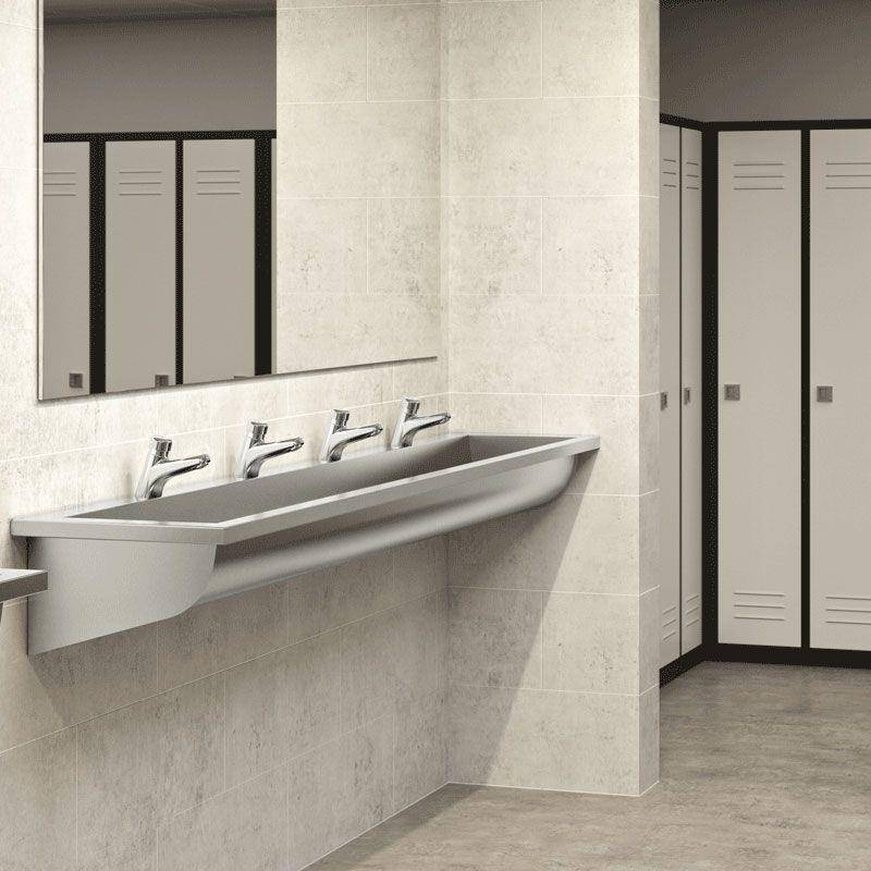 Modernes öffentliches Badezimmer mit einem langen wandmontierten Waschbecken, vier Wasserhähnen, Spiegel und Spindschranktüren an einem gekachelten Innenraum.