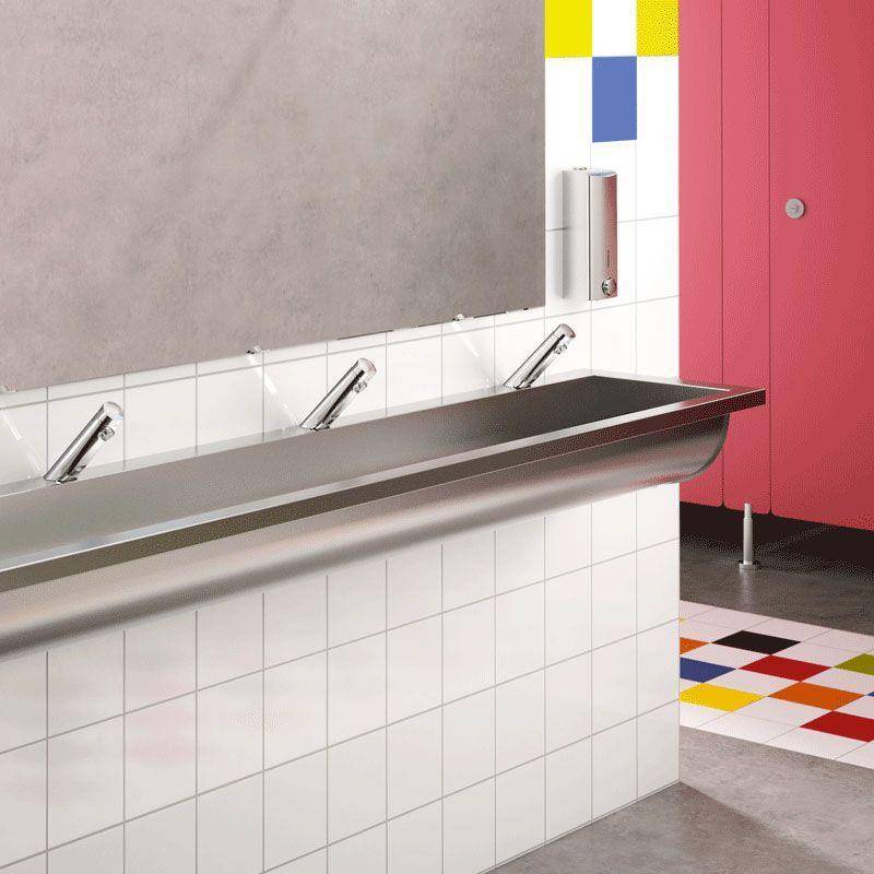 Modernes Badezimmer mit Edelstahlwaschbecken und wandmontierten Wasserhähnen entlang einer gekachelten Ablage, neben rosafarbenen Kabinentüren und einem farbig gefliesten Boden.