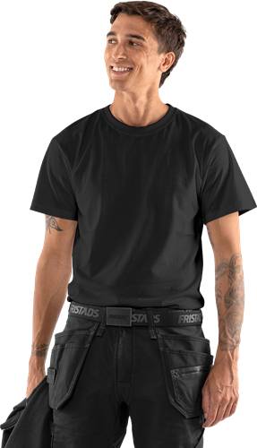 Eine Person trägt ein schwarzes T-Shirt und schwarze Hose mit der Aufschrift 'FRISTADS' am Bund. Ihre Arme sind tätowiert.