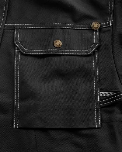 Eine Nahaufnahme einer schwarzen Stoffschürze mit einer großen vorderen Tasche und Messingknöpfen mit weißen Nähdetails.