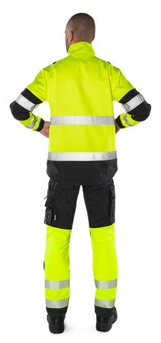 Eine Person trägt eine Warnschutzweste mit reflektierenden Streifen und steht mit den Händen in den Hüften. Die Kleidung umfasst eine dunkle Hose.