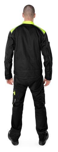 Eine Person in einem schwarzen Outfit steht mit dem Rücken zum Betrachter, die Hände an den Seiten; auf der Hose ist ein grünes Muster sichtbar.