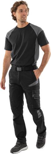 Eine Person trägt schwarze Einsatzhose und ein schwarzes Hemd und steht mit einer Hand in der Tasche.