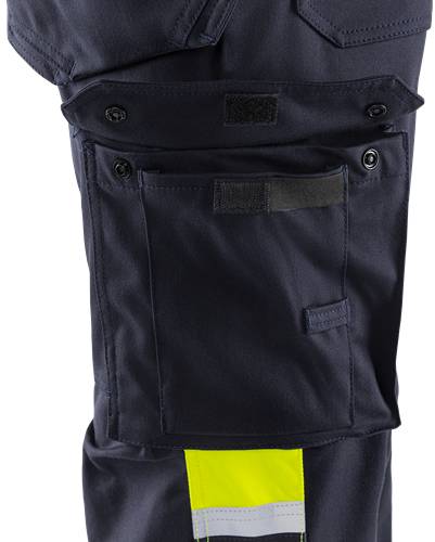 Nahaufnahme einer dunklen Tasche an Kleidungsstück mit einer Klettverschluss-Klappe, die einen kleinen gelben Reflektorstreifen am unteren Rand aufweist.