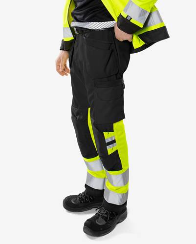 Eine Person trägt hochsichtbare Arbeitshose und Sicherheitsschuhe. Die Hose ist schwarz mit reflektierenden gelben Streifen und mehreren Taschen.