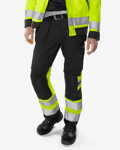 Eine Person, die schwarze und neongelbe reflektierende Arbeitskleidung trägt, bestehend aus Hose und Jacke, die vor einem weißen Hintergrund steht.