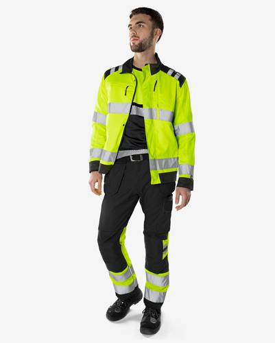 Eine Person, die eine hochsichtbare gelbe Jacke und schwarze Hose mit reflektierenden Streifen trägt und vor einem einfarbigen Hintergrund steht.