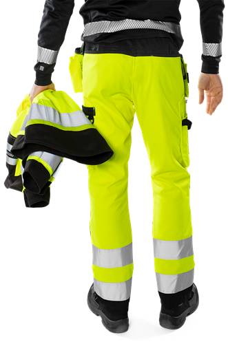 Person trägt eine neongelbe, reflektierende Arbeitshose und hält eine passende Jacke, wobei Sicherheitskleidung hervorgehoben wird.