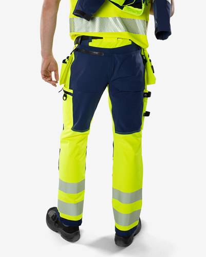 Eine Person trägt Arbeitsschutzhosen mit Reflektionsstreifen, hält ein Werkzeug in einer Hand und steht vor einem grauen Hintergrund.